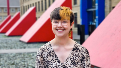 A white mid twenties person stands smiling outside in front of some large red triangle structures. They are wearing a leopard print low cut v-neck top and round glasses. They have brown hair with a thick stripe of bleached hair down the left hand side.