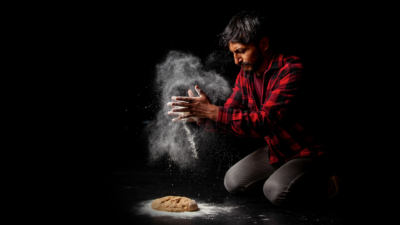 An image of a Bolivian man in a red checked shirt and black jeans spreading flour over some dough on the floor. He is brightly lit against a black background.