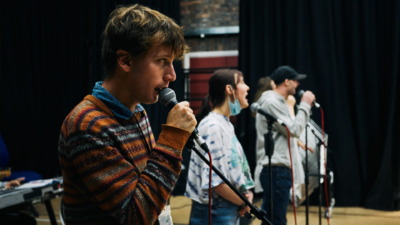 A side shot of a row of men and women singing into microphones in a studio. The person in focus is a white man in his mid twenties with floppy blonde hair and is wearing a striped jumper.
