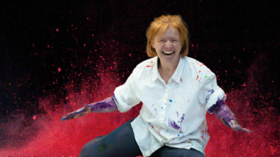 A red haired white woman in her thirties is sat on the floor throwing red powder paint in the air. She is wearing a white smock and her arms are covered in purple paint