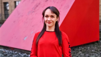 A headshot of Sarah Chapelo. Sarah is a white woman with chest-length, straight brown hair. She is wearing a red, long sleeved top. She is smiling. The photo is taken outside, at dusk, in front of some large red triangular vents.