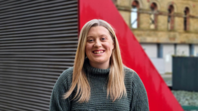 A headshot of Emily Blackwell. Emily is a white woman with chest-length, straight blonde hair. She has blue eyes and is smiling, showing cheek dimples and teeth. She is wearing a dark mossy green knitted jumper. The photo is taken outside in front of large red triangular vents.