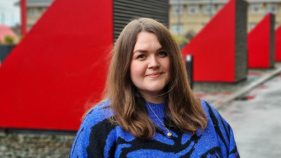 A headshot of Jess Boyes. Jess is a white woman with slightly wavy, shoulder length medium brown hair. She is wearing a blue and black zebra print jumper with a shiny gold locket necklace. The photo is taken outside, in front of large red triangular vents.