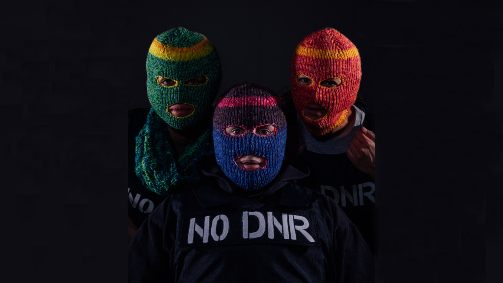 Three people stare directly at the camera and are each wearing different coloured, knitted balaclavas where only their eyes and mouths are exposed. They are all wearing black stab vests with the words ‘NO DNR’ stencilled onto the vests in white paint. Charmaine is on the left of the image. She is wearing a knitted green balaclava and blue knitted snood. Chopin is on the right of the image. She is wearing a red and orange knitted balaclava. She is tugging the top of the stab vest with her left hand. Jess is in the centre of the image between Charmaine and Chopin and is staring straight at the camera. She is wearing a blue and pink knitted balaclava.