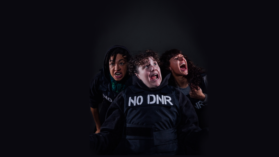 Three people shout in different directions with varying exaggerated expressions of rage, confusion and anticipation. They are all wearing black stab vests with the words ‘NO DNR’ stencilled onto the vests in white paint. Jess, a white woman with curly brown hair is in the centre of the image. Her mouth is open and she is looking upwards as she shouts. Charmaine, a light skinned black woman of mixed heritage, is behind Jess to the left of the image. She is wearing a green knitted snood and is looking into the distance with an expression of extreme frustration. Chopin, a white person with long dark brown curly hair and a fringe. She has both of her eyes closed and her mouth is open as she shouts with her head tilted upwards.