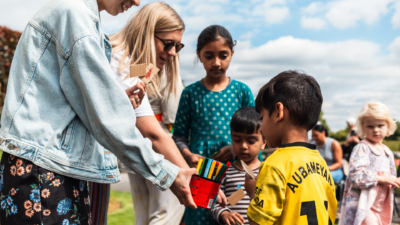 A woman with blonde hair and wearing sun glasses is holding a box of pens to a child.