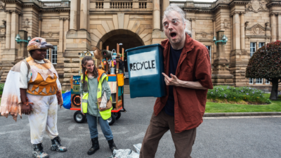 In front of a grand stone building three performers stand in front of a cart. This mobile home carries many home objects such as a shower, an oil can, a robot and a doll house. It has colours of brown, yellow and blue. In front of the cart two performers are facing each other. Closest to the camera, a white man with grey hair, wearing a brown shirt and trousers is holding a blue bin that says Recycle. The are pointing towards the bin as they look to the right with a very expressive expression on their face. To the left of him, stands two female performers. In the middle stands Homie wearing a bright yellow visor jacket, blue trousers and black boots. On the left stands Birdie, wearing a brown bird hat, white and brown winds, a brown plumage vest and black boots. They are looking towards the recycling bin. In front of them on the ground lies a mix of recycling including bottles, electrical items and clothing.