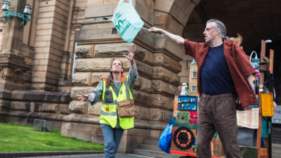 A male performer on the right waring a blue t-shirt and brown shirt is holding a metal litter picker with a plastic bag on the end in the air to the left. Below the plastic bag a female performer wearing a yellow high-vis jacket is reaching towards the plastic bag.