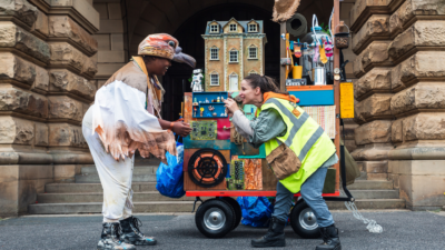 Performance of Birdie. A travelling cart is behind two performers. This mobile home carries many home objects such as a shower, an oil can, a robot and a doll house. It has colours of brown, yellow and blue. In front of the cart two performers are facing each other. On the left, a young female is wearing a brown bird had, white and brown wings and black boots. They have their hands in front of them and are leaning towards Homie. Homie, a young female, is wearing a bright yellow visor jacket, blue trousers and black boots. They are holding something in their hands and leaning towards Birdie.
