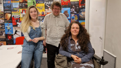 A photo of Paul Wilshaw, Melissa Johns and Cherylee Houston smiling at the camera. On the left Melissa stands with blonde hair, wearing blue dungarees and brown boots. Next to her stands Paul with brown curly hair, a white shirt and blue jeans. In front in a wheelchair sits Cherylee wearing a blue zip up hoodie, a blue and black skirt and white shoes. They are standing in a room with posters of theatre shows on the wall behind them.