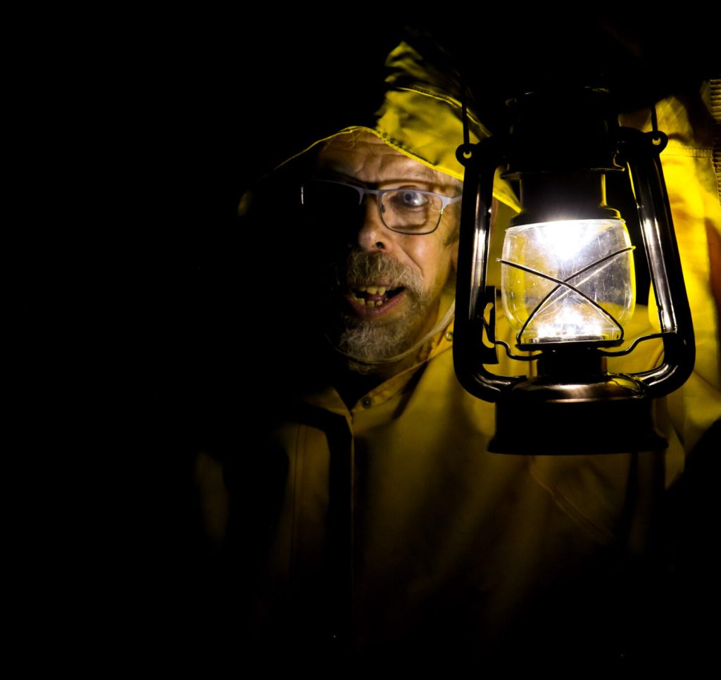 Jez, a man with glasses and a beard, is wearing a yellow raincoat with the hood up and his holding an old fashioned lantern towards us